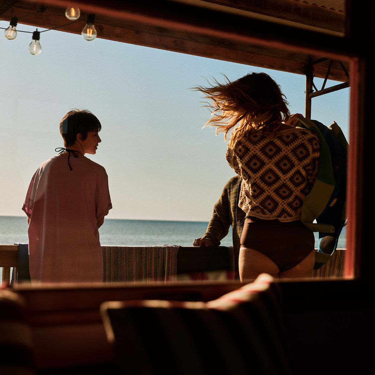 Two women with surfboards on the front deck of a holiday house overlooking the ocean.