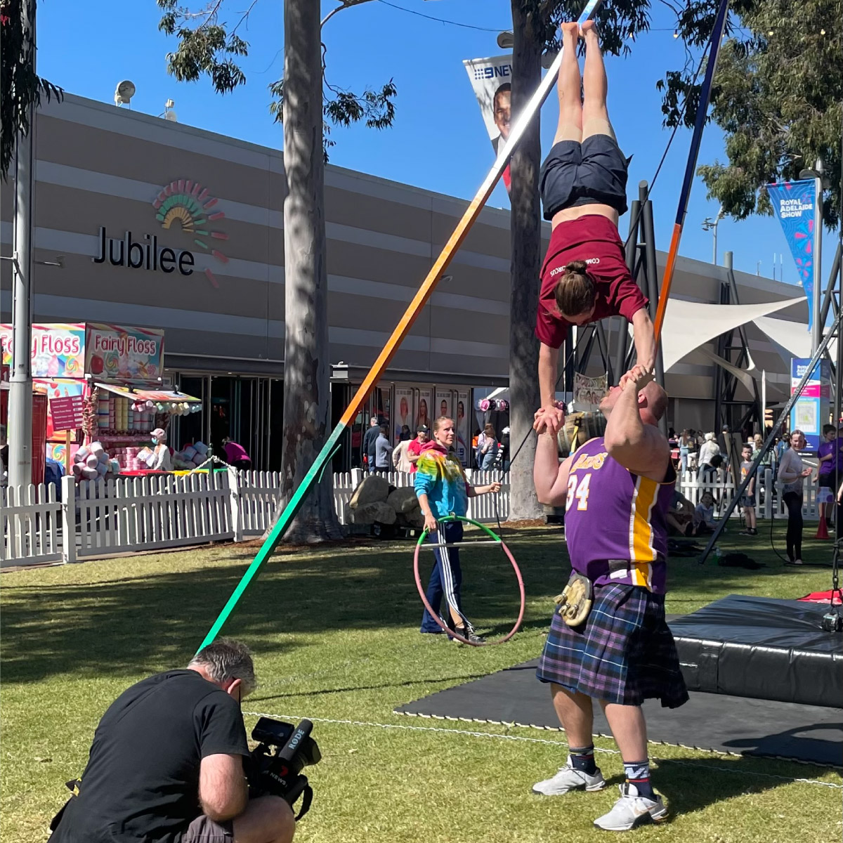 Acrobats performing in the open show grounds while film crews capture footage.